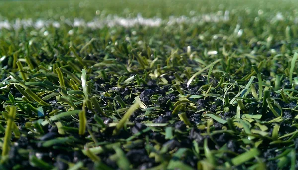 Close up of black rubber-crumb granules packed between the green fibres of a synthetic pitch, a white line behind