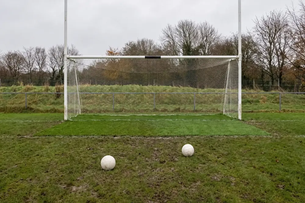 A muddy grass pitch in winter, the goalmouth still green in front of a Gaelic goal, two footballs on the mud